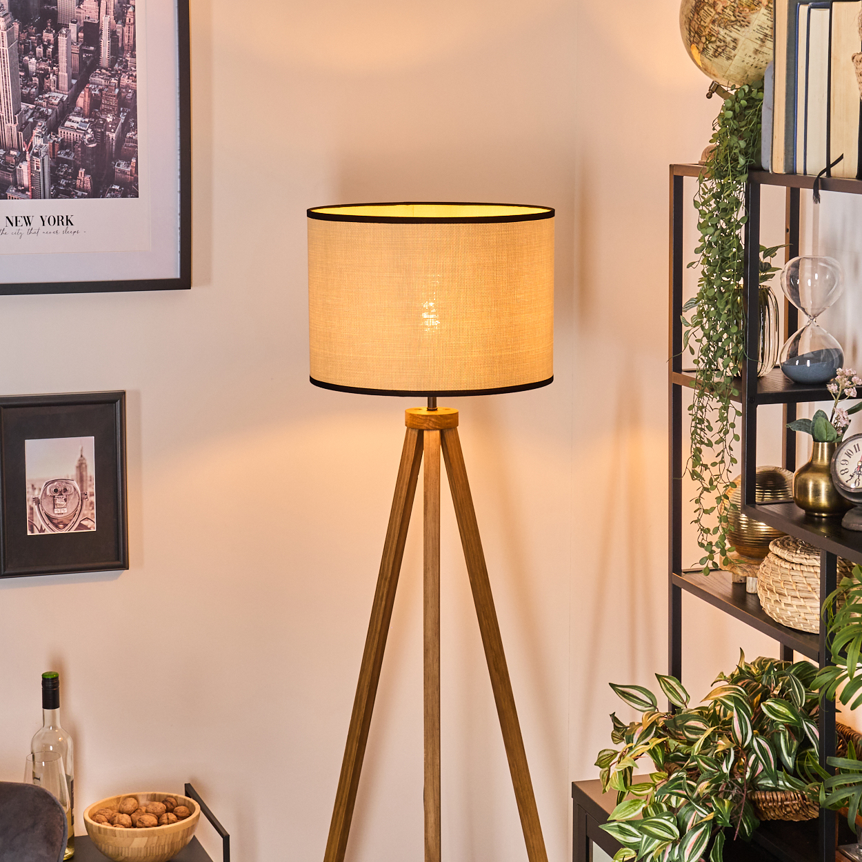 Eine Holz-Stativlampe mit beigem Schirm leuchtet. Regale im Hintergrund haben Bücher und Pflanzen. Es gibt ein New York-Bild mit Rahmen.