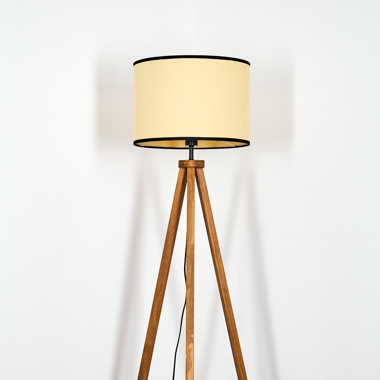 Eine hölzerne Stehlampe mit Stativ steht vor einer weißen Wand. Die Lampe hat einen cremefarbenen Lampenschirm mit schwarzem Rand.