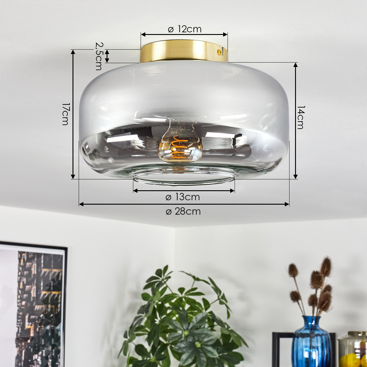 Eine moderne Deckenlampe mit Maßen. Glasschirm mit Goldaufsatz. Eine Glühbirne im Inneren. Im Hintergrund Pflanzen und Dekoration.