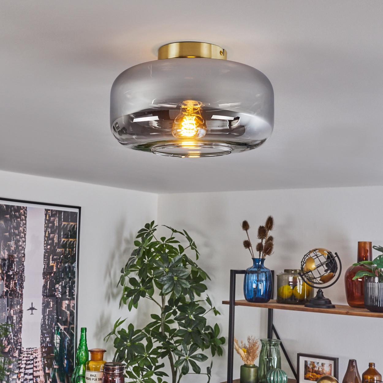 Eine moderne Deckenlampe mit leuchtender Glühbirne. Darunter ein Bücherregal mit Deko und einer Pflanze. Ein Stadtbild links.