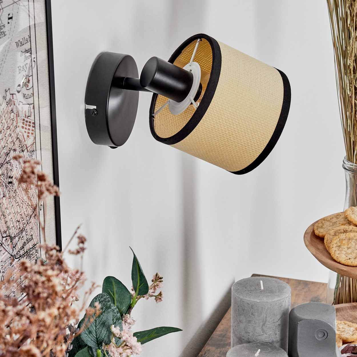 Eine schwarze Wandlampe mit zylindrischem beigen Schirm hängt an einer weißen Wand. Auf einem Holztisch stehen Kerzen, Kekse und getrocknete Pflanzen.