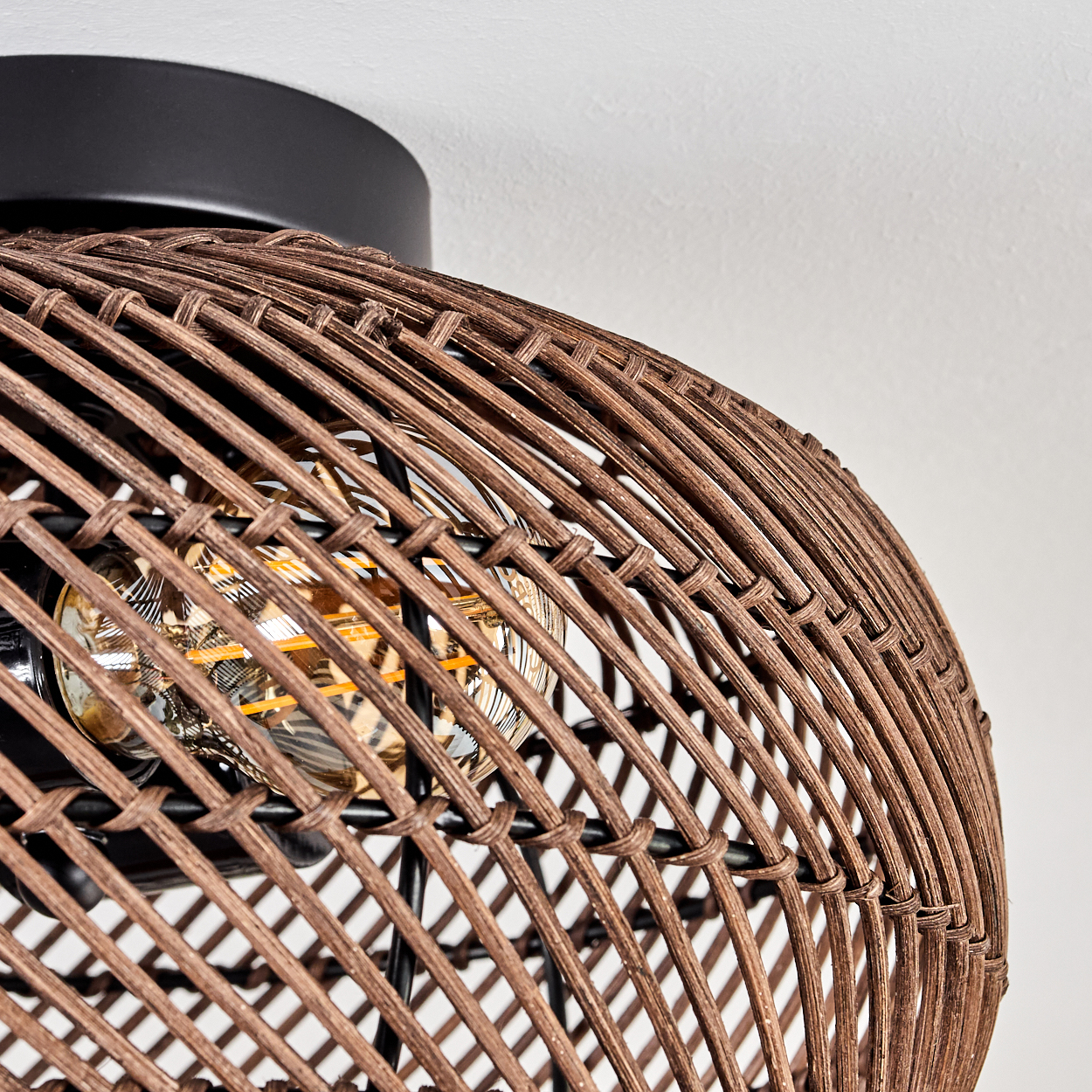 Nahaufnahme einer braunen, gewebten Rattanlampe mit sichtbarer Glühbirne. Die Lampe ist vor einem weißen Hintergrund und einem schwarzen Sockel.