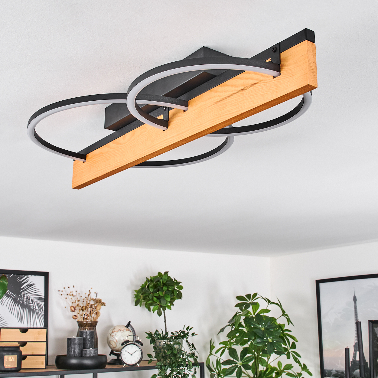 Eine moderne Deckenlampe mit Holzbalken und schwarzen Metallringen. Pflanzen und Dekoration sind unter der Lampe sichtbar.