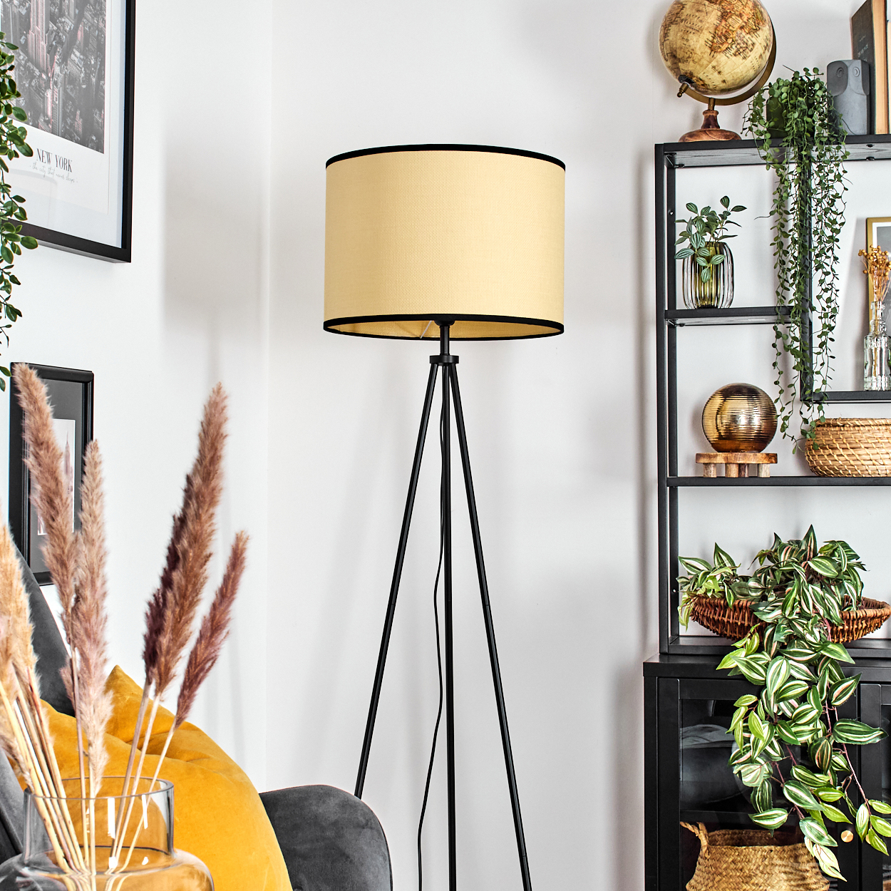Ein Zimmer mit Stehlampe, Regal mit Dekorationen und Pflanzen sowie Sofa mit Kissen. Die Lampe hat einen beigefarbenen Lampenschirm und schwarze Beine.