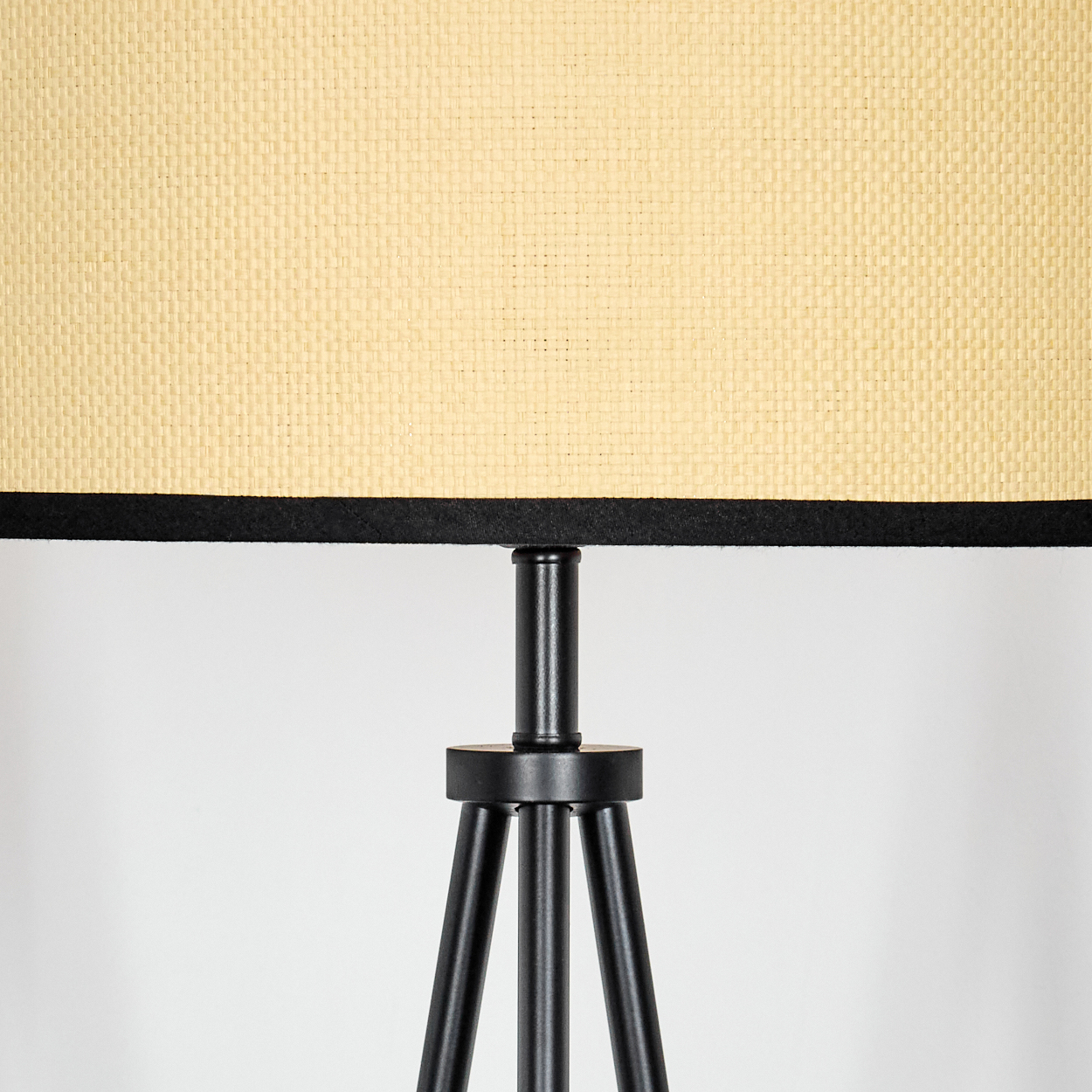 Eine Lampe mit beigem gewebtem Schirm und schwarzer Umrandung. Die Lampe hat ein schwarzes Metall-Dreibein vor weißem Hintergrund.