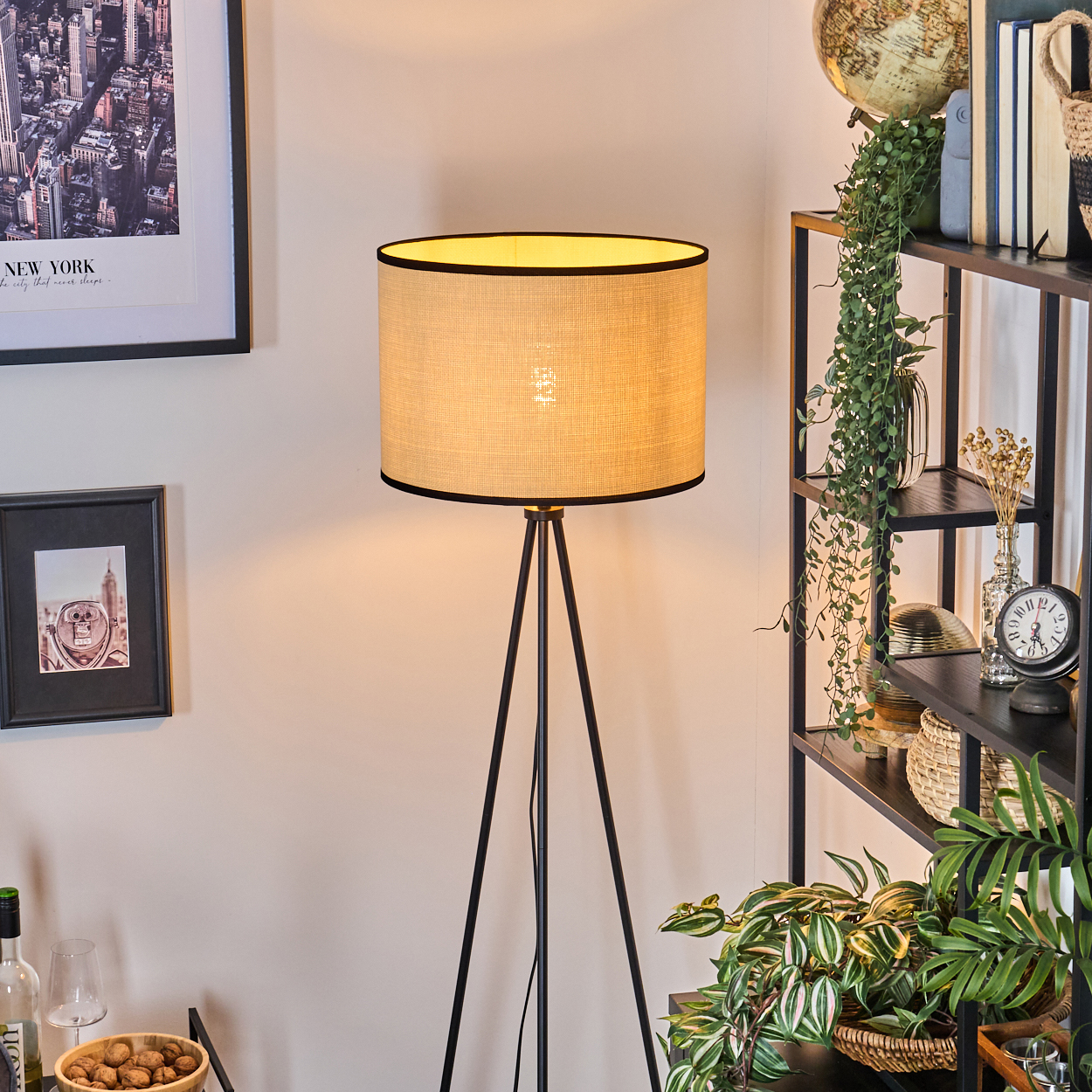 Eine Stehlampe mit beigem Schirm beleuchtet einen Raum mit Bücherregal und gerahmten Bildern.
