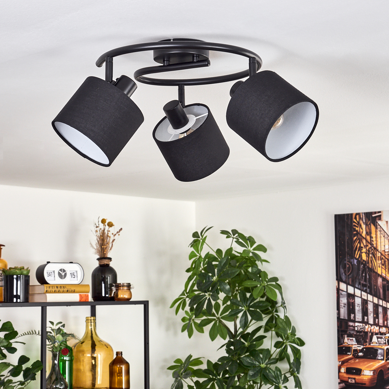 Eine schwarze Deckenleuchte mit drei verstellbaren Lampen. Darunter sind eine Pflanze und Dekoration zu sehen. Das Licht ist zentriert.