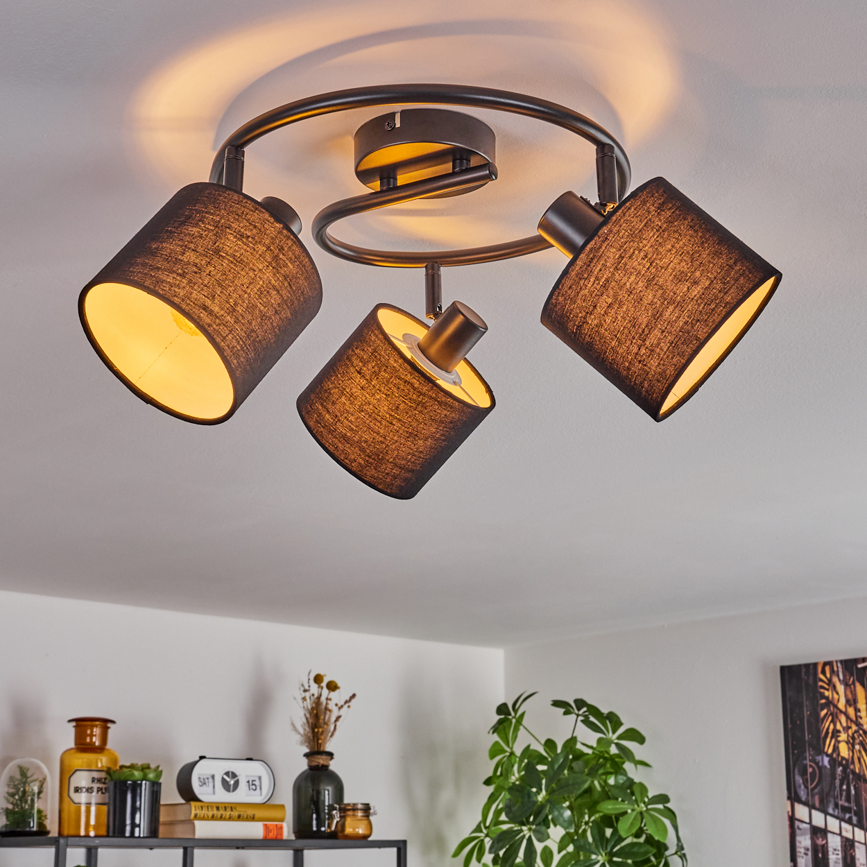 Eine Deckenleuchte mit drei schwarzen Lampenschirmen. Die Lampen sind nach unten geneigt und beleuchten den Raum. Pflanzen und Deko sind unten sichtbar.