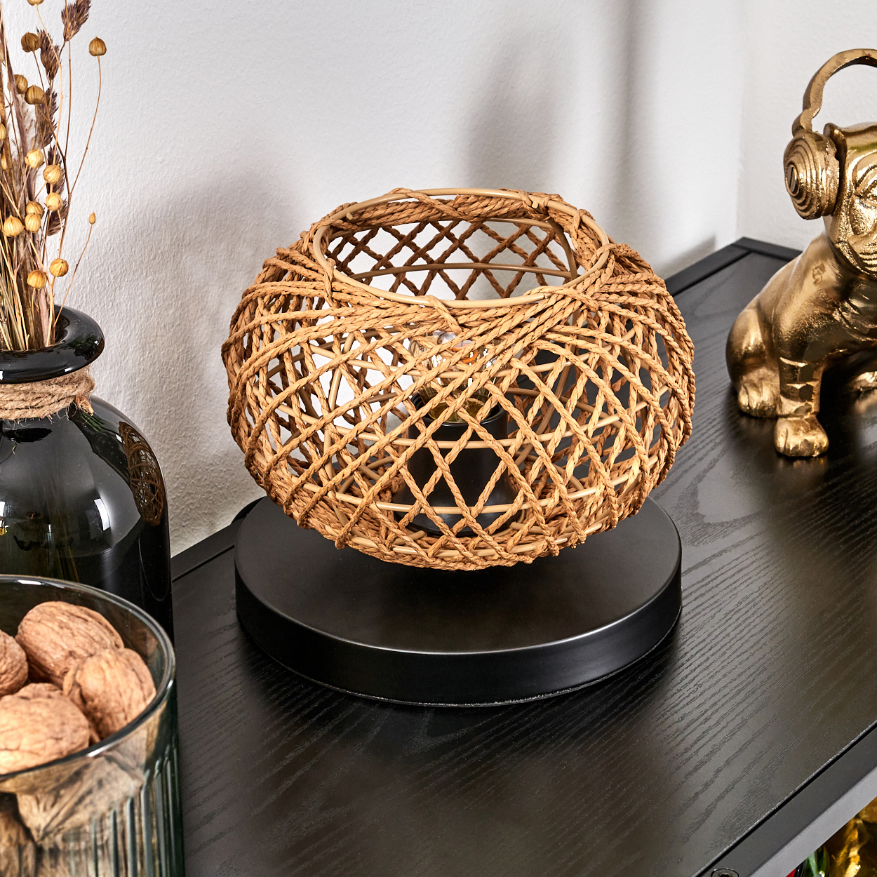 Eine gewebte Lampe steht auf einem schwarzen Tisch. Rechts steht eine goldene Hundestatue mit Kopfhörern. Links eine Vase mit trockenen Pflanzen.