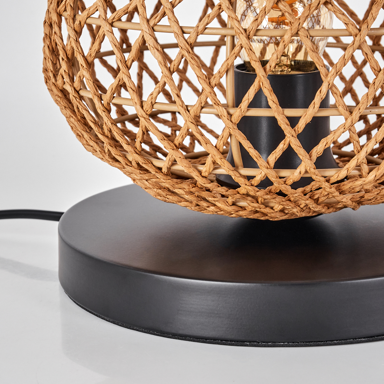 Nahaufnahme einer Lampe. Sie hat einen gewebten Rattanschatten, einen schwarzen Sockel und eine sichtbare Glühbirne. Der Hintergrund ist weiß.