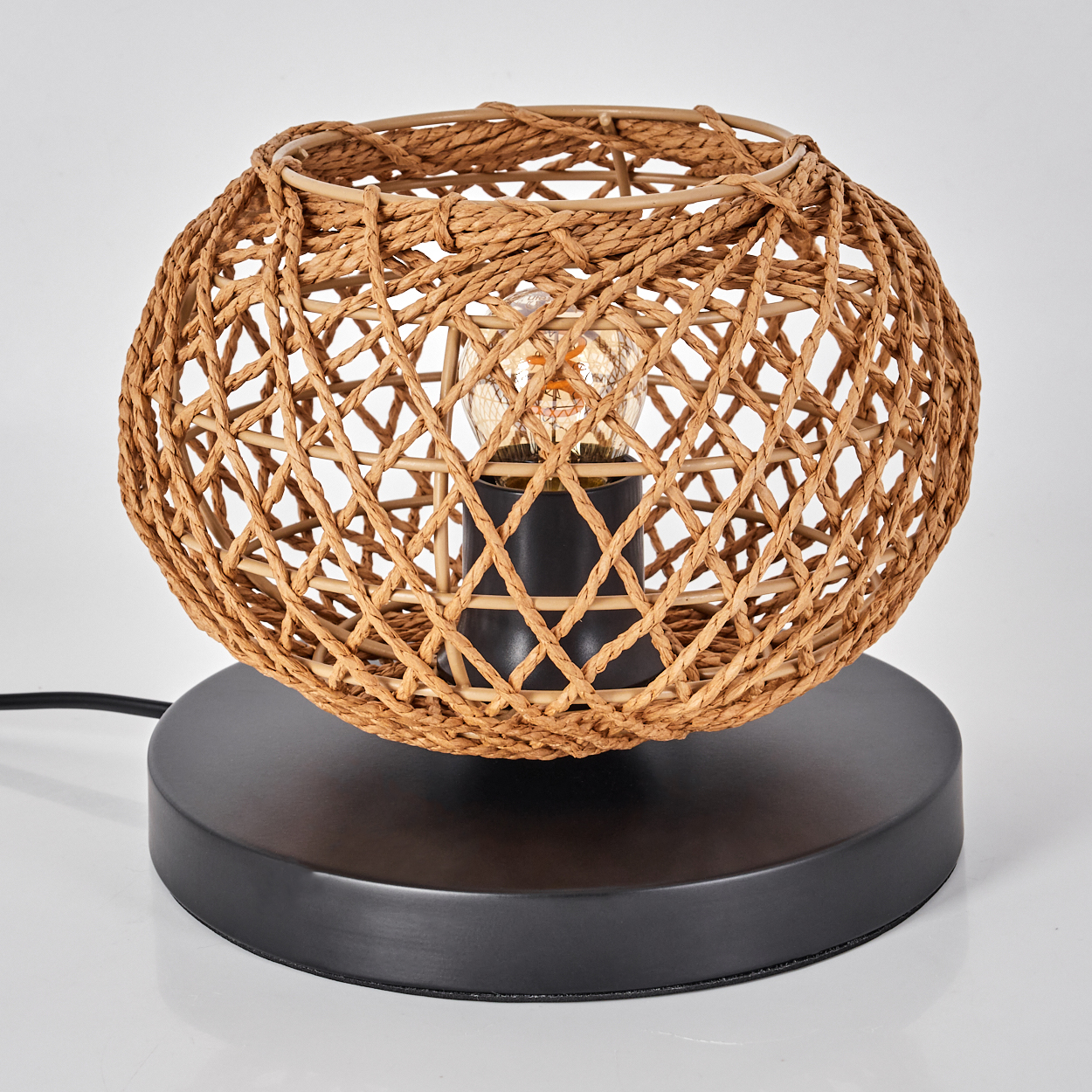 Eine gewebte Korblampe mit schwarzem Sockel und freiliegender Glühbirne. Die Lampe steht auf einer weißen Oberfläche.