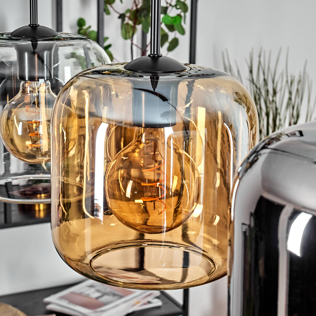 Nahaufnahme von drei Pendelleuchten mit bernsteinfarbenen Glasschirmen und sichtbaren Glühbirnen. Hintergrund: Pflanzen und Tisch.