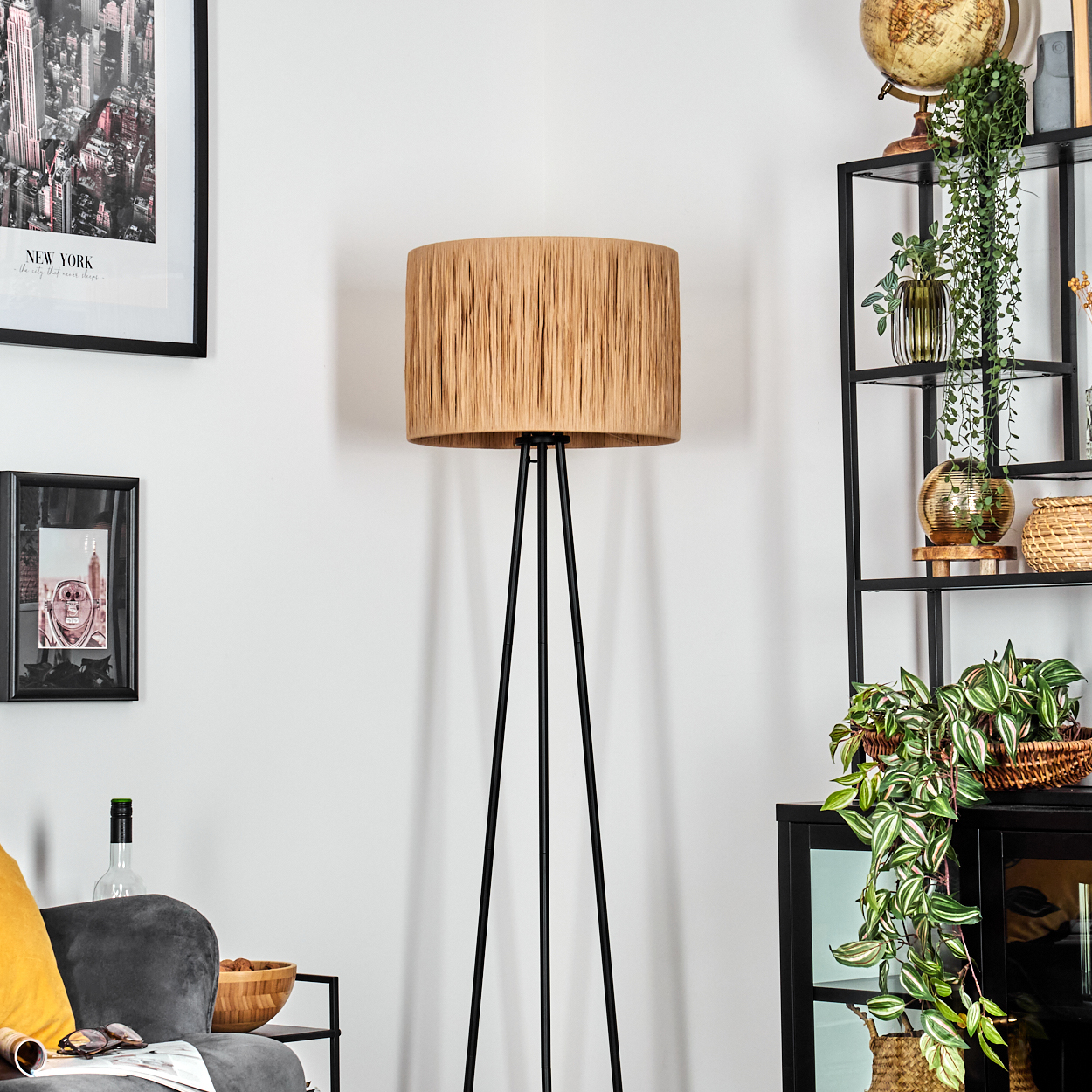 Eine Stehlampe mit beigem Lampenschirm steht in einem Raum. An der Wand befindet sich ein Regal mit Pflanzen und einem New York-Bild.