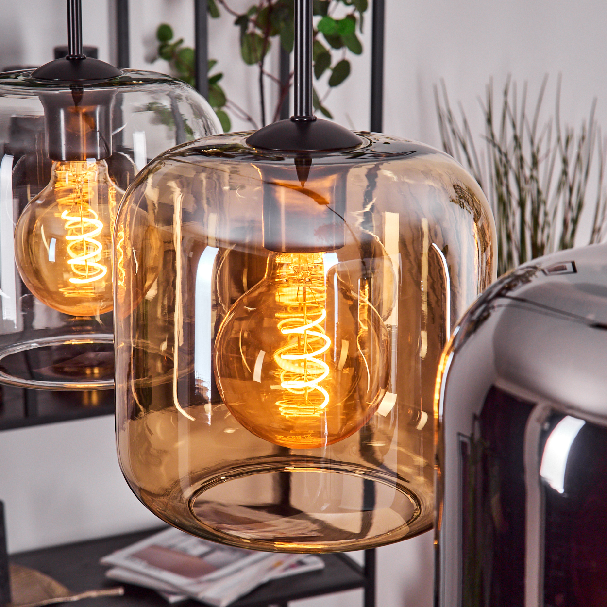 Nahaufnahme von drei hängenden Pendelleuchten. Glasschirme mit bernsteinfarbener Farbe, je mit einer Wendelglühbirne. Hintergrund hat Pflanzen.