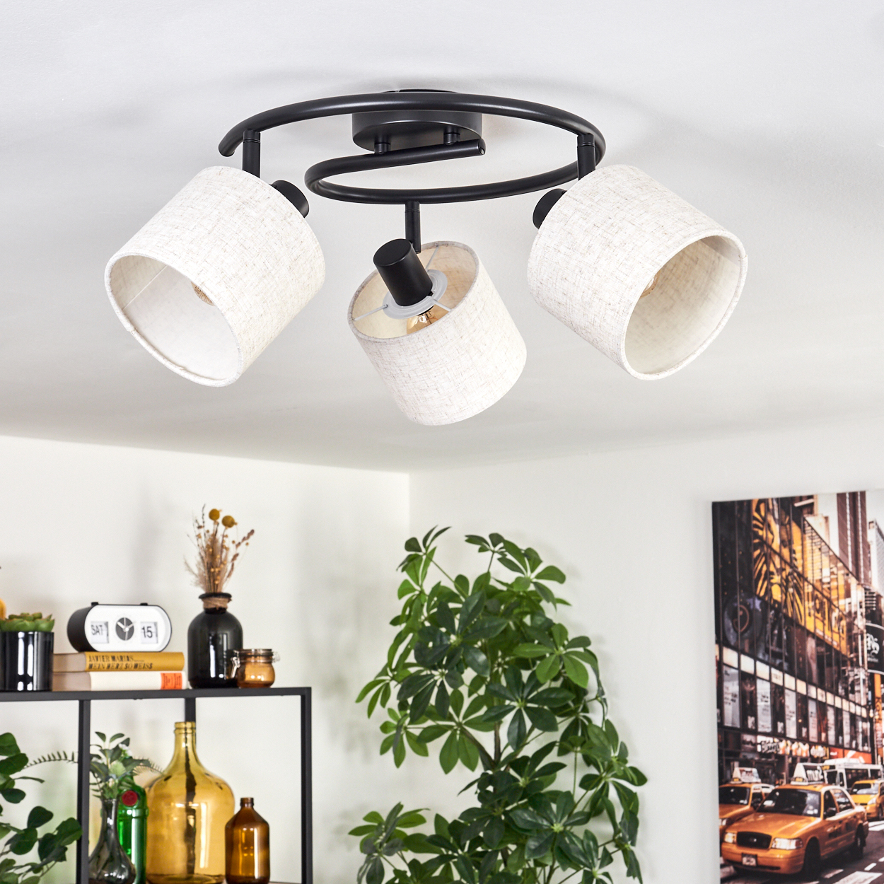Eine moderne Deckenlampe mit drei beigefarbenen Schirmen ist an einer weißen Decke montiert. Ein Regal mit Pflanzen und Vasen ist sichtbar.