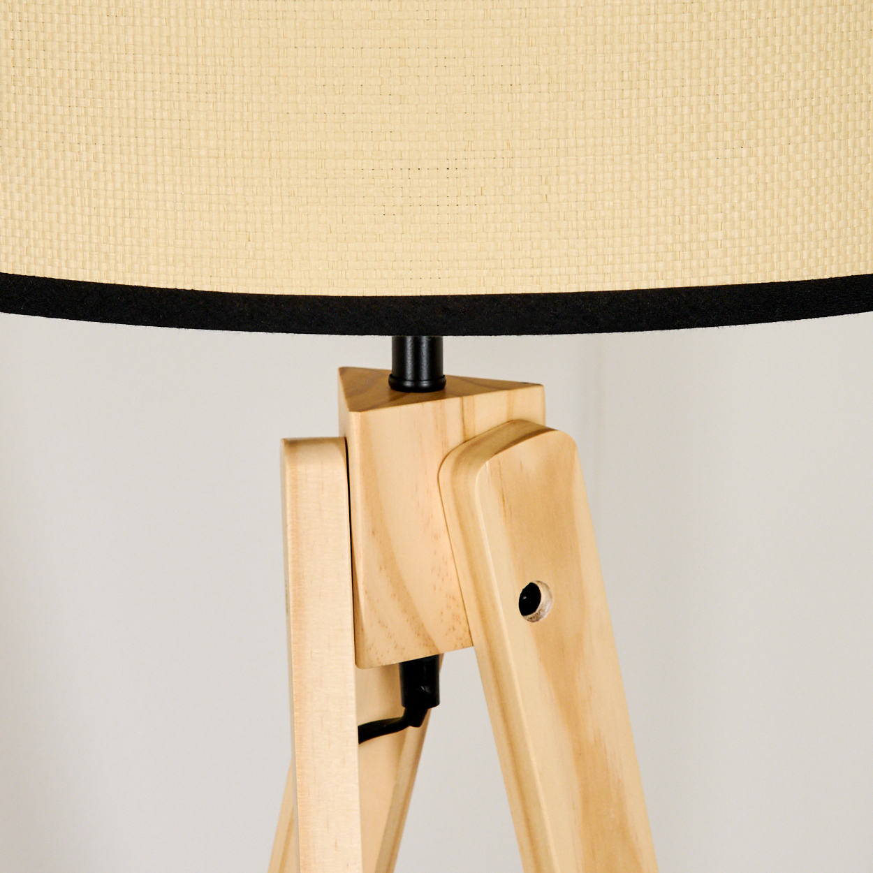 Eine Holz-Stativlampe mit beigem Lampenschirm. Die Lampe hat eine schwarze Einfassung und einen schwarzen Stützfuß. Der Hintergrund ist eine hellgraue Wand.