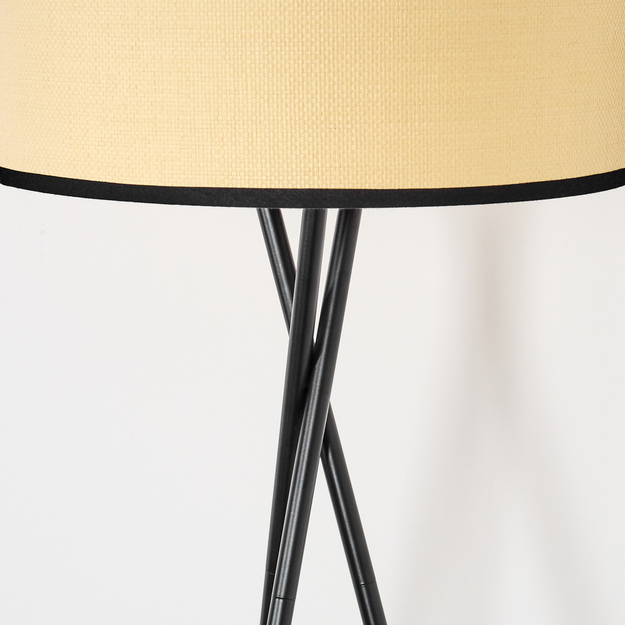 Eine Stehlampe mit beigem Lampenschirm und schwarzem Rand. Der Sockel ist schwarz und hat drei Beine. Der Hintergrund ist weiß.