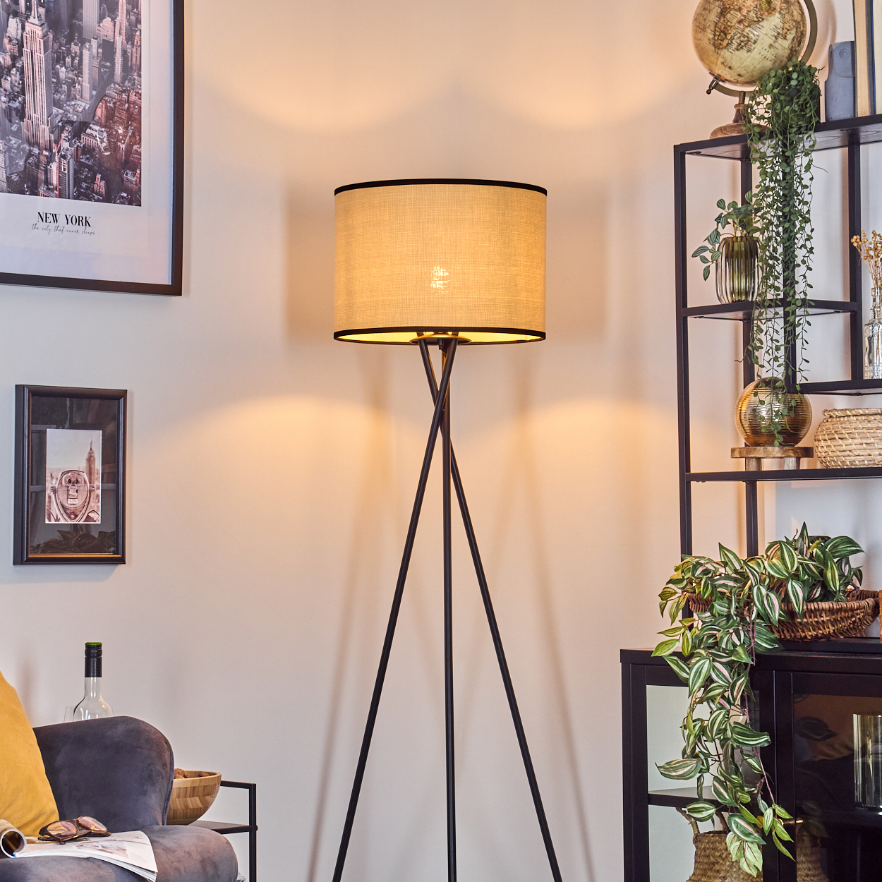 Eine beleuchtete Lampe steht in einem Raum. Rechts steht ein Regal mit Dekoration. Kunstwerke hängen an der Wand. Ein Sofa ist sichtbar.