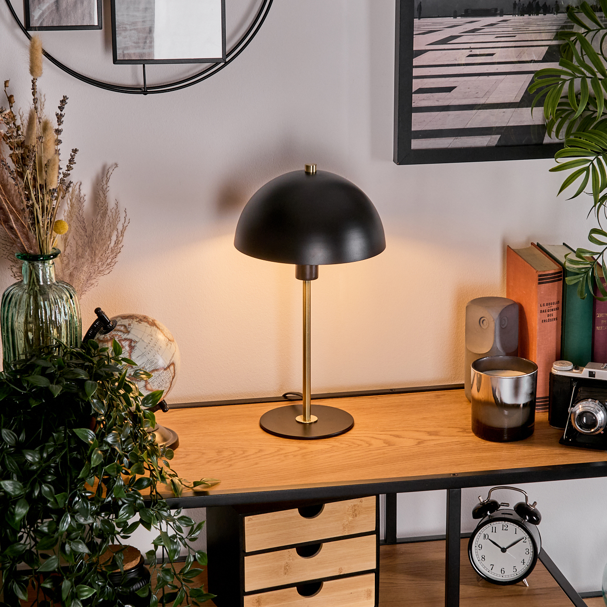 Eine schwarze Lampe auf einem Holztisch mit Büchern, einem Globus, einer Uhr und Pflanzen. Hintergrund: gerahmte Kunst und eine Wand.