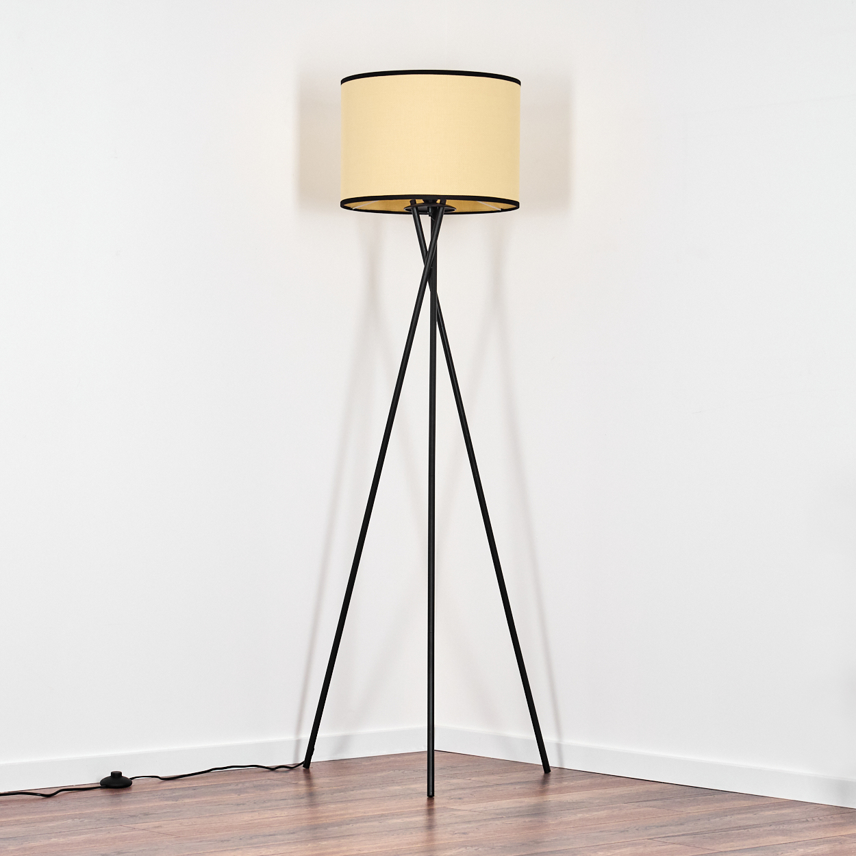 Eine schwarze Stativ-Stehlampe mit beigem Lampenschirm steht in einer Ecke. Der Boden ist aus Holz, die Wände sind weiß.