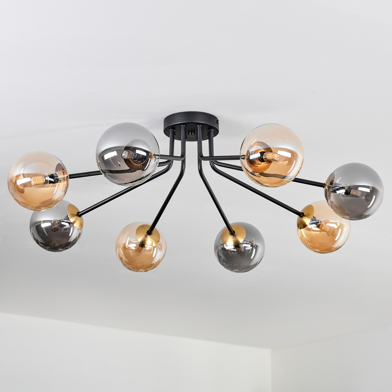 Eine moderne Deckenleuchte mit acht kugelförmigen Lampen. Die Glühbirnen sind in Gold und Silber. Der Rahmen ist schwarz, vor einer weißen Decke.
