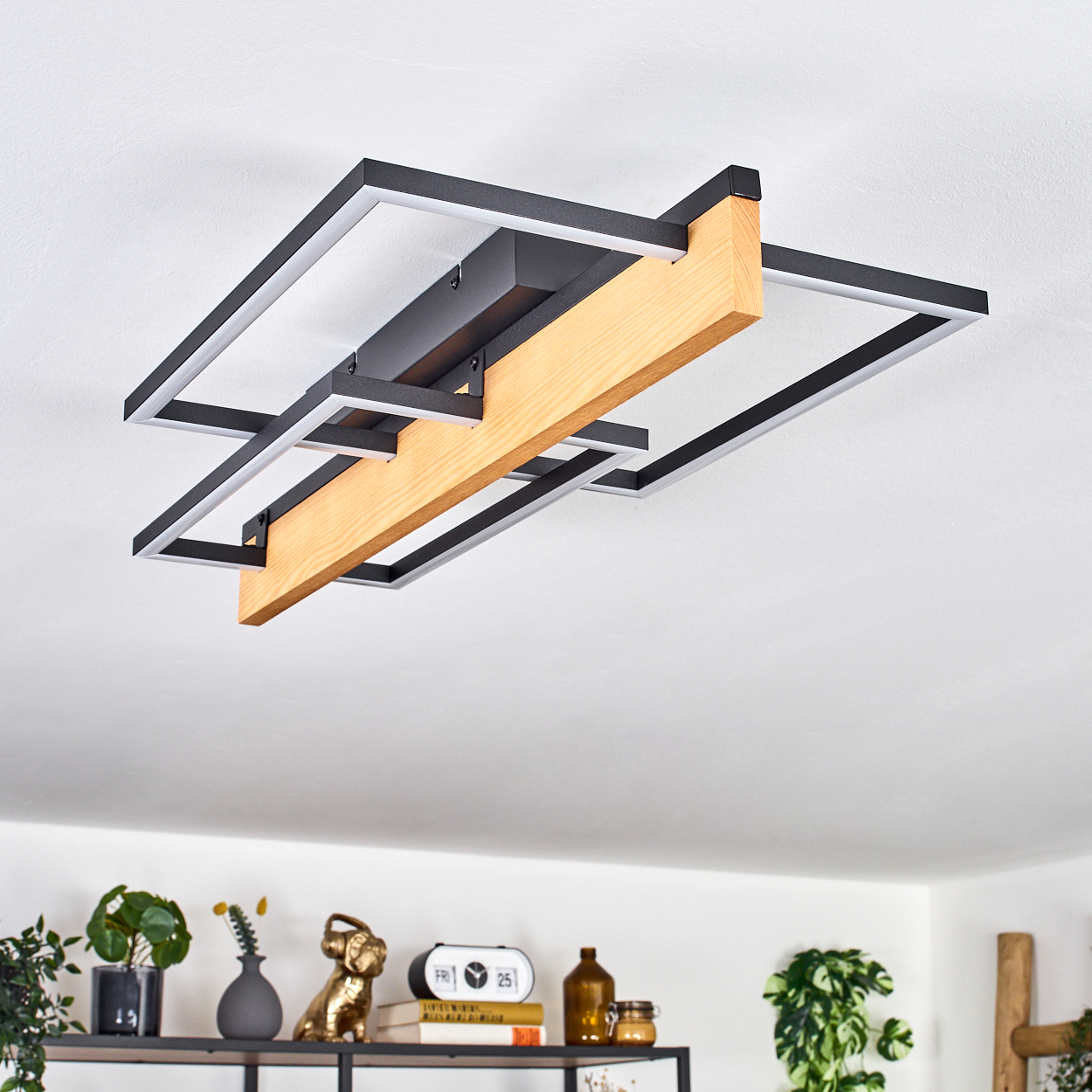 Eine moderne Deckenlampe mit schwarzen Rahmen, weißen Lichtern und einem Holzbalken. Darunter ein Regal mit Pflanzen, Dekor und Büchern.