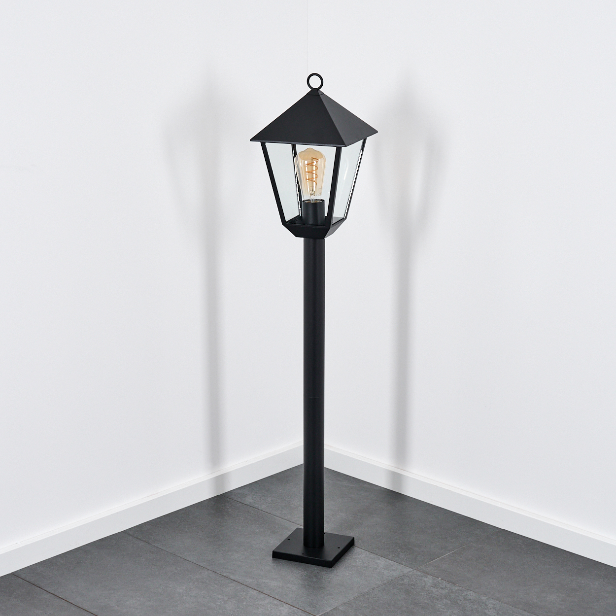 Eine schwarze Außenlampe steht in einer Ecke. Sie hat eine Glühbirne und steht auf einem quadratischen Sockel.