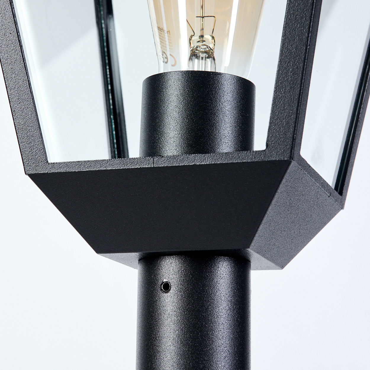 Nahaufnahme einer schwarzen Außenlampe. Sie hat einen zylindrischen Sockel, einen geometrischen Rahmen mit Glasscheiben und eine Glühbirne.
