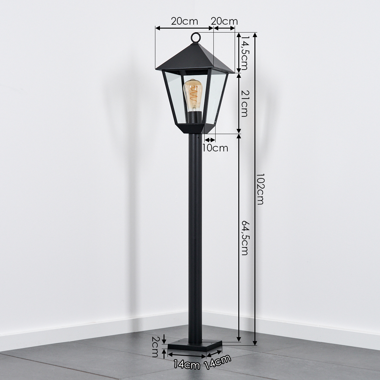 Eine schwarze Außenlampe mit angegebenen Abmessungen. Sie hat ein Glasgehäuse und eine sichtbare Glühbirne, vor einer weißen Wand und grauem Boden.