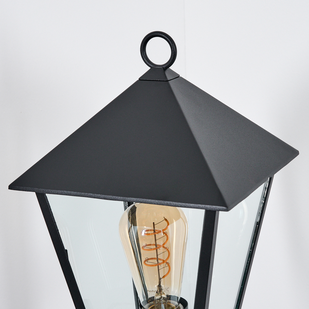 Nahaufnahme einer schwarzen Außenlampe mit Glasgehäuse. Im Inneren befindet sich eine Glühbirne im Vintage-Stil. Der Hintergrund ist eine weiße Wand.