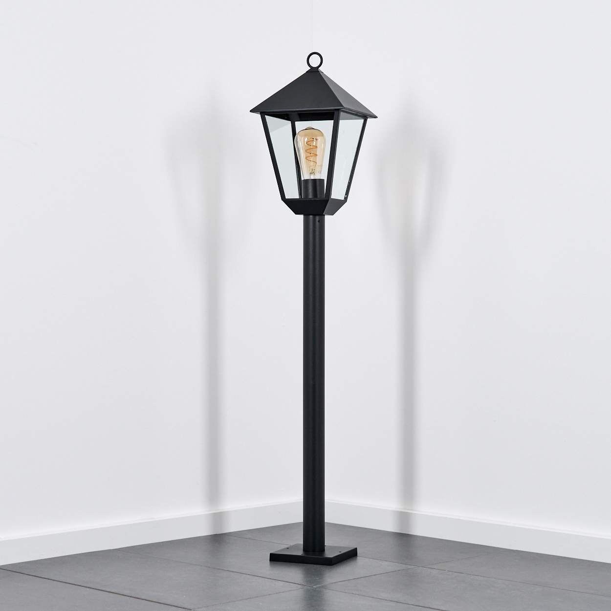 Eine schwarze Außenlampe steht in einer Ecke. Sie hat ein Glasgehäuse mit einer Glühbirne, einen hohen Pfahl und eine quadratische Basis.
