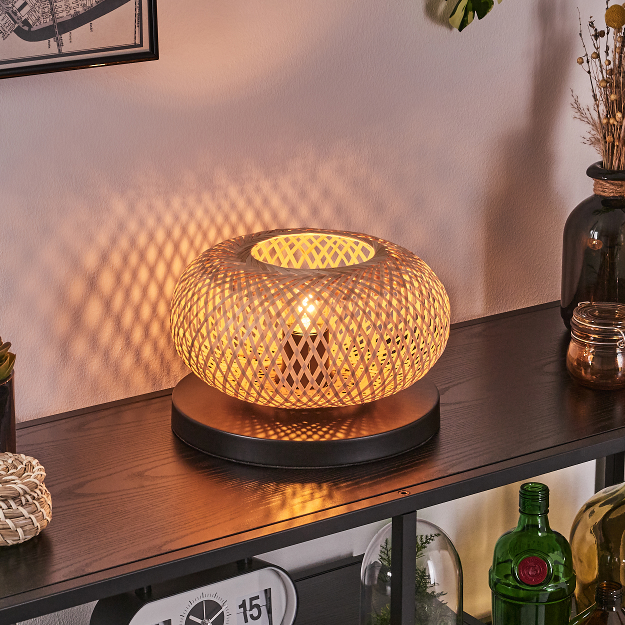 Eine gewebte Rattanlampe auf einem Regal mit anderen dekorativen Gegenständen wie Vasen, Pflanzen und einer Uhr. Warmes Licht und Schatten.