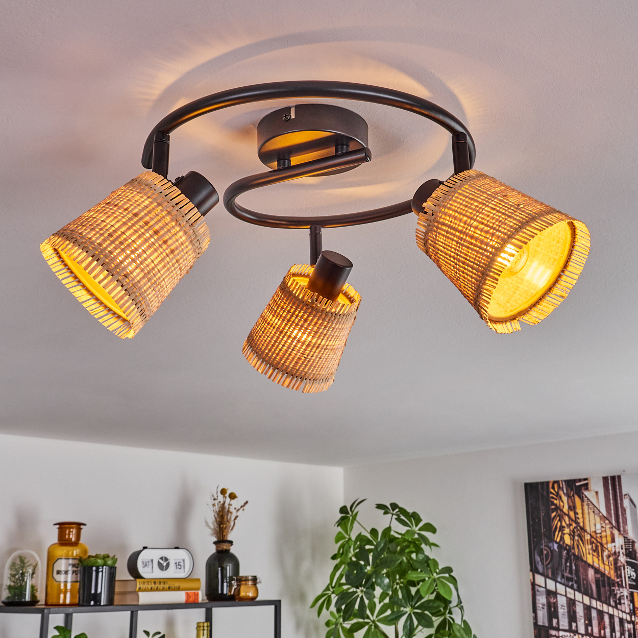 Eine Deckenleuchte mit drei Korblampenschirmen. Die Lampen sind an. Darunter ein Regal mit Dekorationen und eine grüne Pflanze.