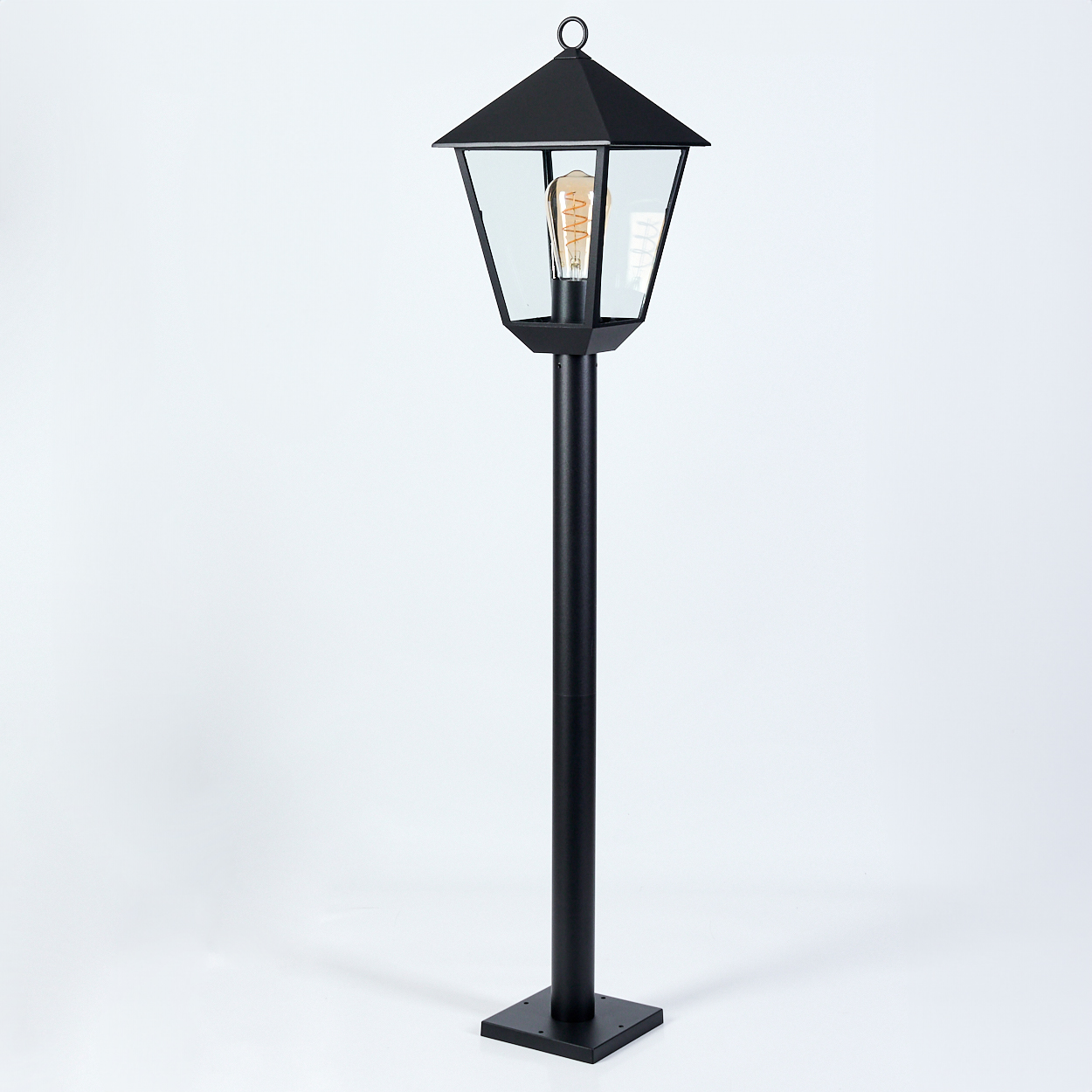 Eine schwarze Außenlaterne. Sie hat einen quadratischen Sockel, eine lange Stange und einen Kopf mit Glasscheiben und einer Glühbirne.
