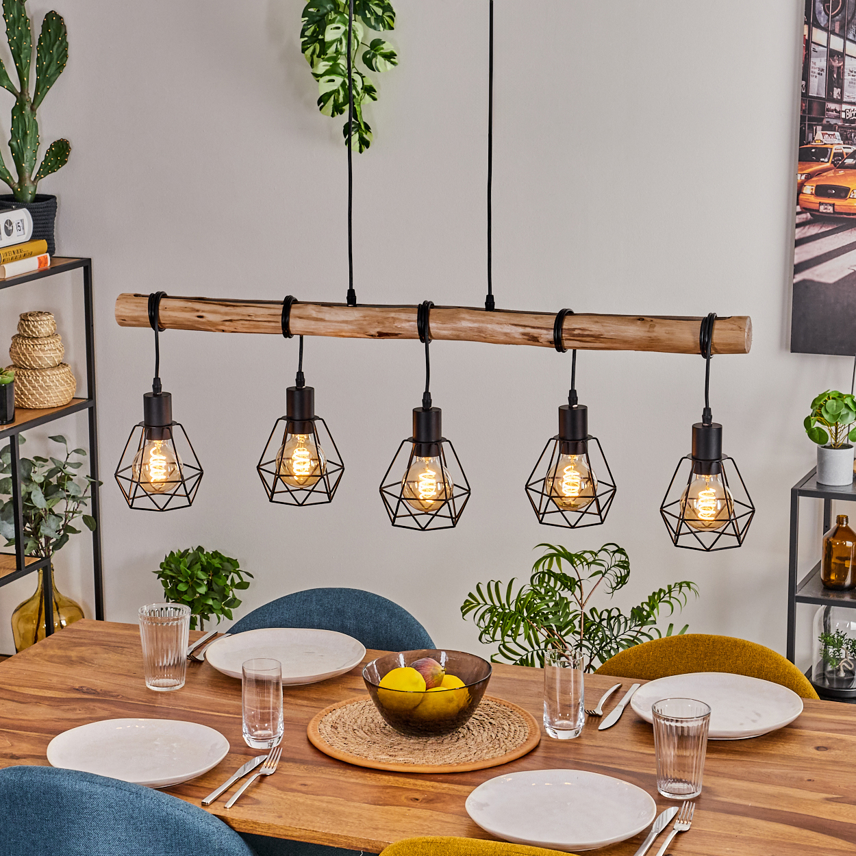 Esszimmer mit Holztisch, Stühlen und einer Leuchte mit fünf Glühbirnen. Eine Obstschale und Teller sind gedeckt.