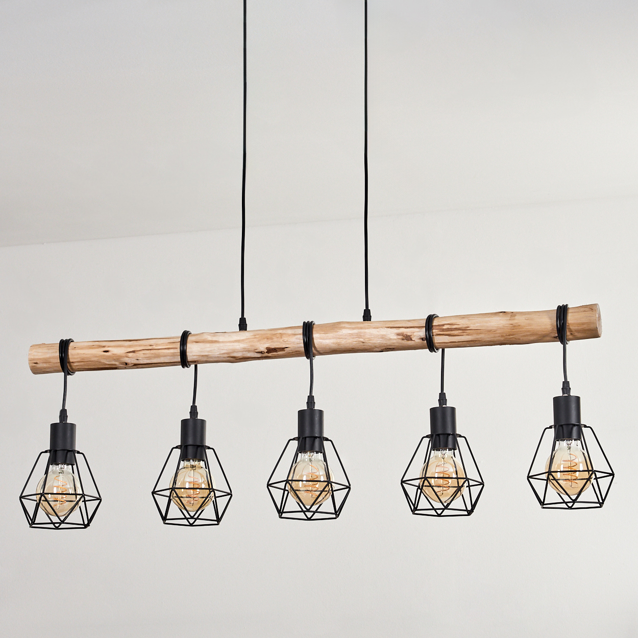 Ein Holzbalken mit fünf schwarzen Pendelleuchten. Jede Leuchte hat einen Drahtkäfig um eine Glühbirne. Der Hintergrund ist eine weiße Wand.