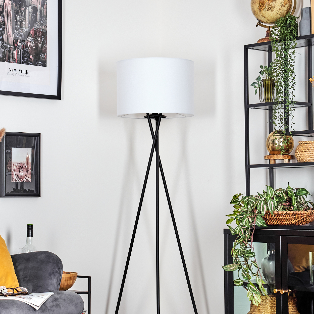 Ein Zimmer mit weißem Lampenschirm auf schwarzen Beinen, ein schwarzes Metallregal mit Pflanzen und ein New York City Poster an der Wand.