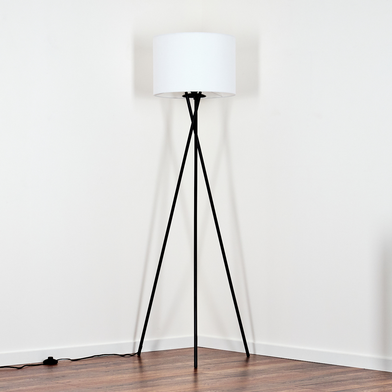 Eine moderne Stehlampe mit weißem Lampenschirm und schwarzen Stativbeinen steht in einer Zimmerecke. Holzboden und weiße Wände.
