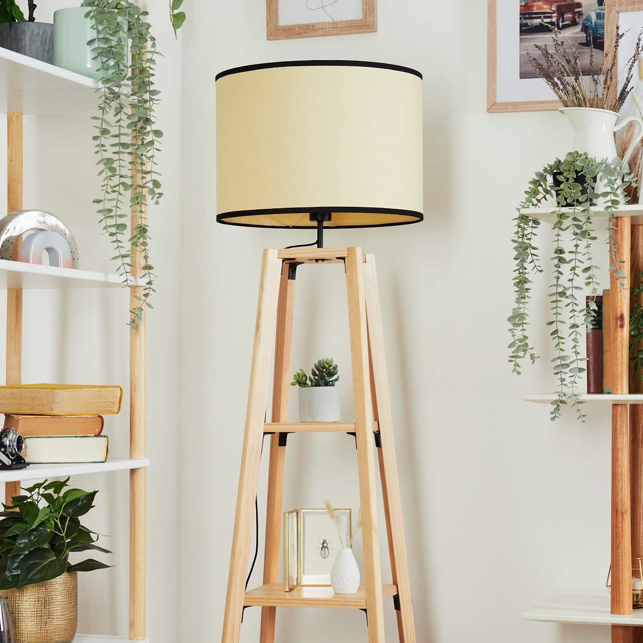 Eine Holzlampe mit cremefarbenem Schirm steht neben einem Bücherregal mit Pflanzen und Dekoration. Ein Bild hängt an der Wand.