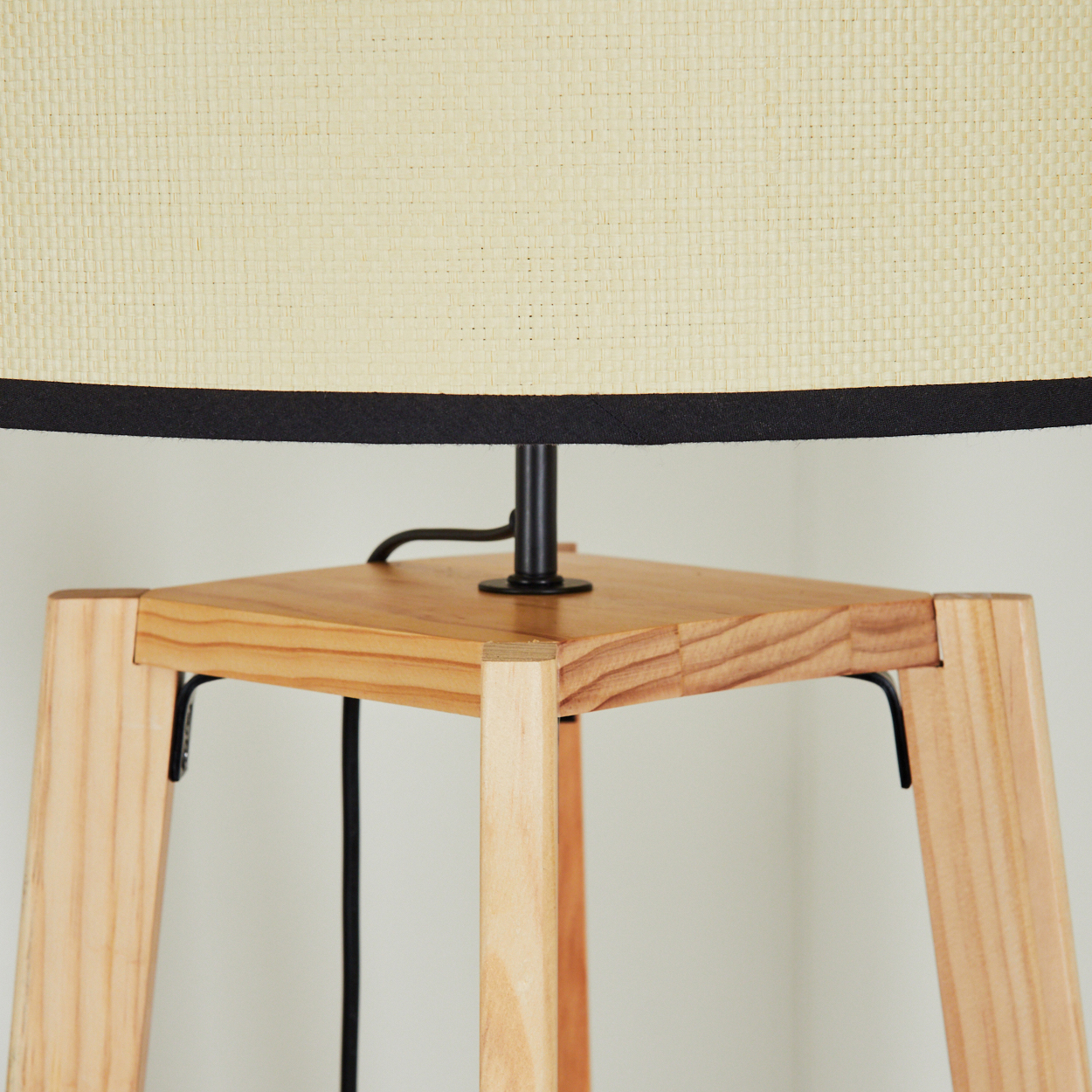 Eine Lampe mit beigem Lampenschirm, schwarzem Rand und Holzsockel. Die Beine sind ebenfalls aus Holz, ein schwarzes Kabel ist sichtbar.