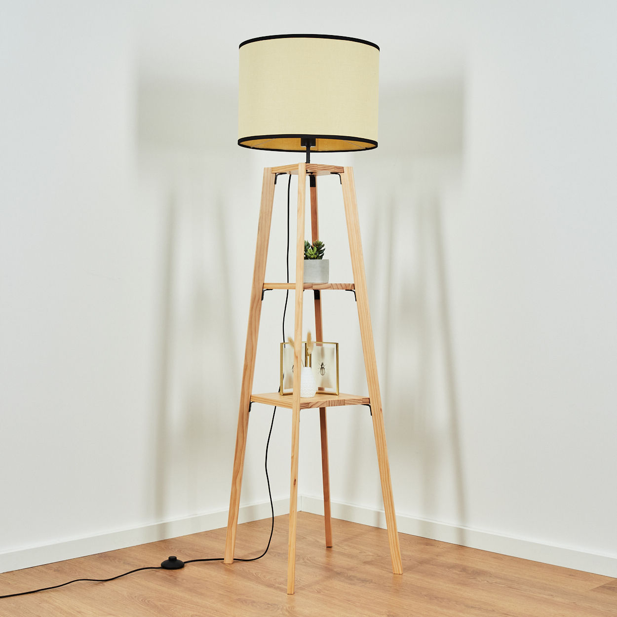 Eine Stehlampe aus Holz mit cremefarbenem Lampenschirm. Sie hat Regale mit einer Pflanze, Bilderrahmen und steht in einer Ecke.