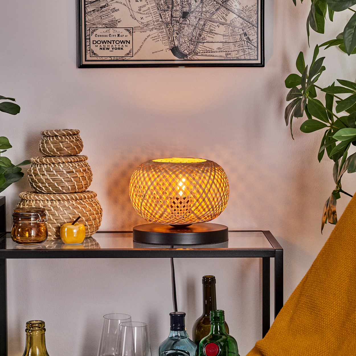 Eine Lampe mit gewebtem Schirm steht auf einem Glastisch. In der Nähe sind gestapelte Körbe, eine Karte, Flaschen und ein gelbes Kissen zu sehen.