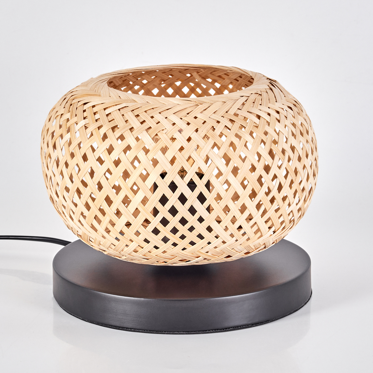 Ein Lampenschirm aus gewebtem Bambus sitzt auf einem dunkelgrauen Sockel. Die Lampe steht auf einer weißen Oberfläche.