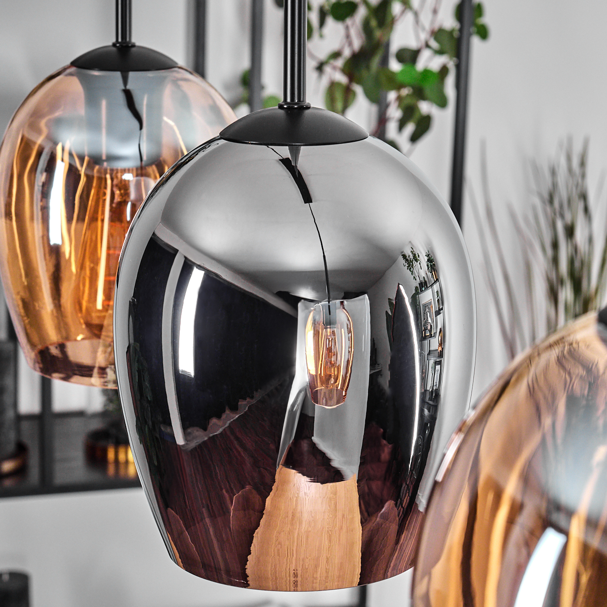Drei kugelförmige Pendelleuchten mit reflektierenden Oberflächen sind aufgehängt. Eine ist chromfarben, andere sind bronzefarben. Hintergrund hat Pflanzen und Möbel.
