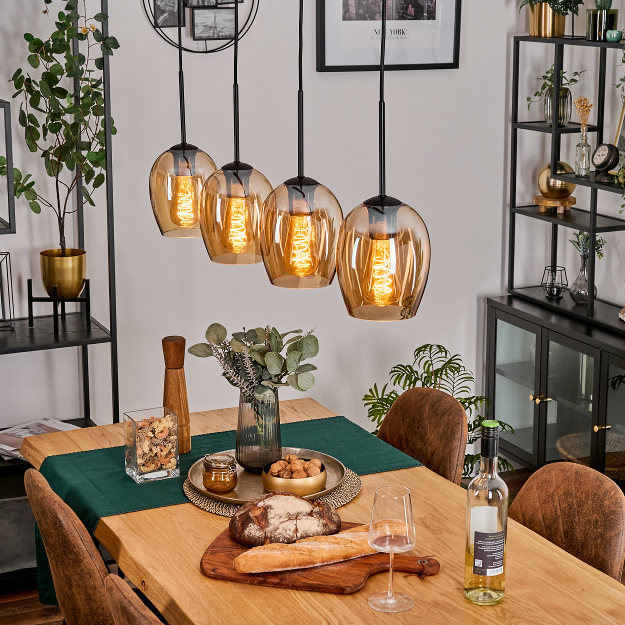 Ein Esstisch mit vier Pendelleuchten darüber. Auf dem Tisch befinden sich Brot, eine Weinflasche, ein Glas und ein Tablett mit Essen.