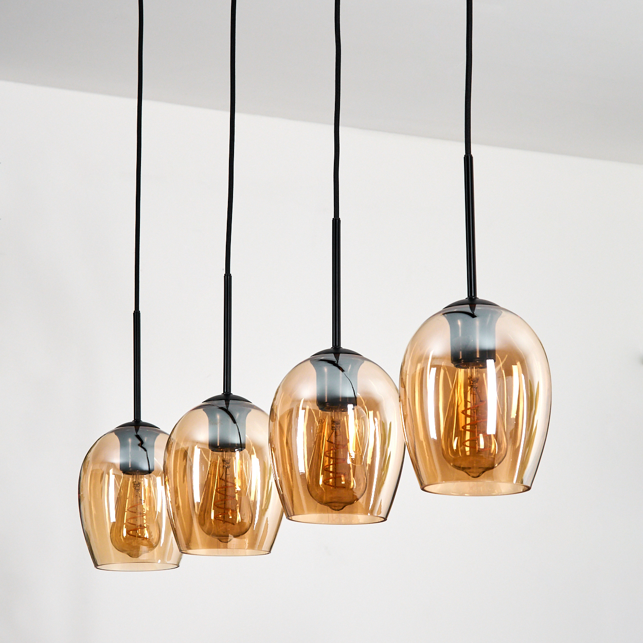 Vier Pendelleuchten mit bernsteinfarbenen Glasschirmen und Edison-Glühbirnen hängen vor weißem Hintergrund, schwarze Kabel sichtbar.