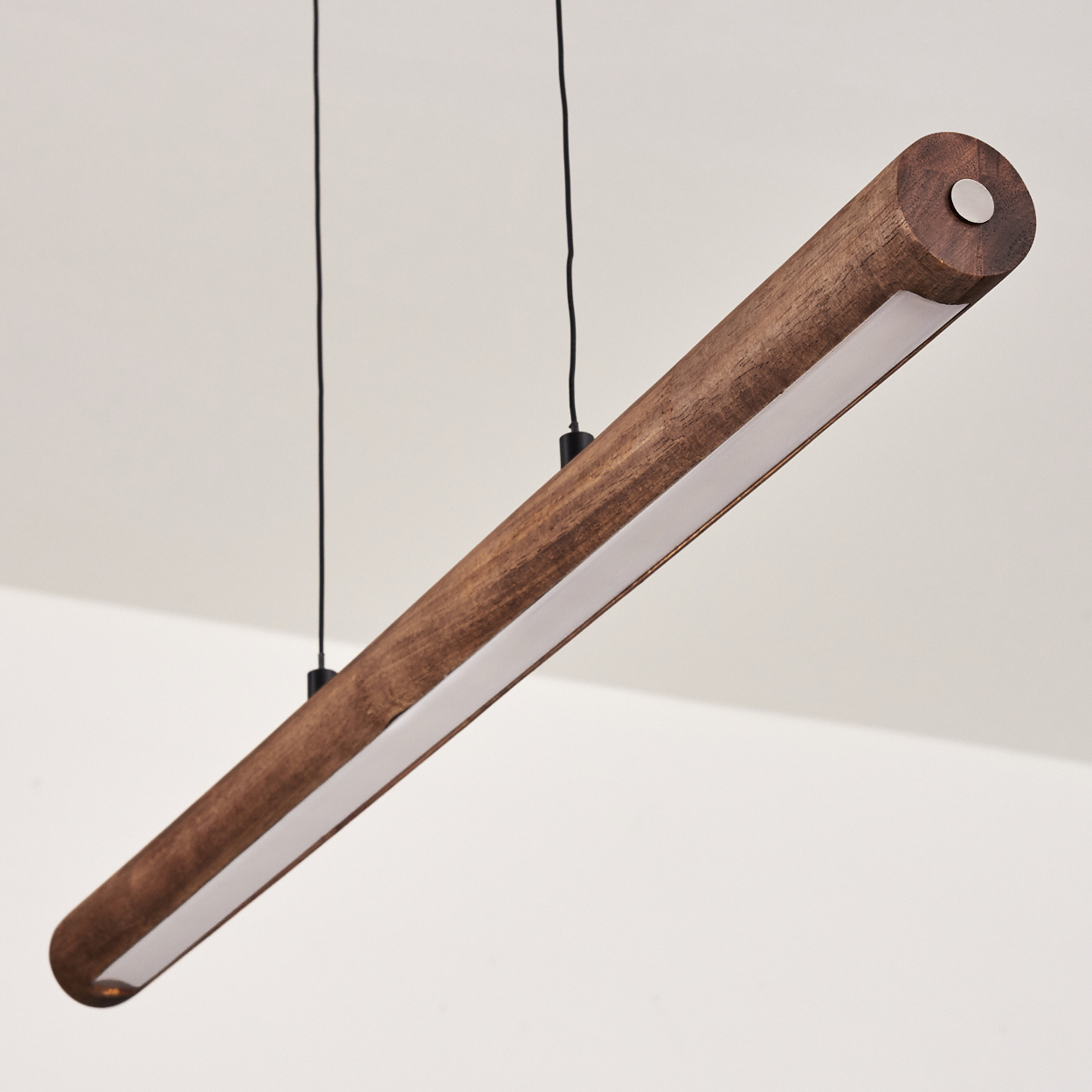 Eine zylindrische Holzleuchte hängt von der Decke. Sie hat einen weißen Lichtstreifen und wird von zwei schwarzen Kabeln gehalten.