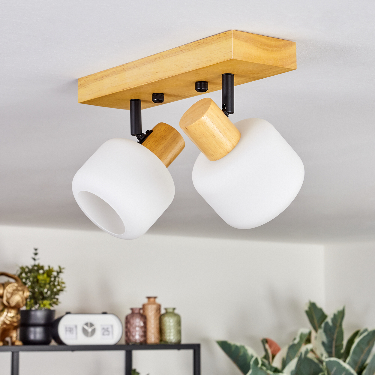 Eine Deckenleuchte mit zwei weißen, abgerundeten Lampen, die an einer Holzbasis befestigt sind. Der Hintergrund umfasst ein Regal mit Dekor und Pflanzen.