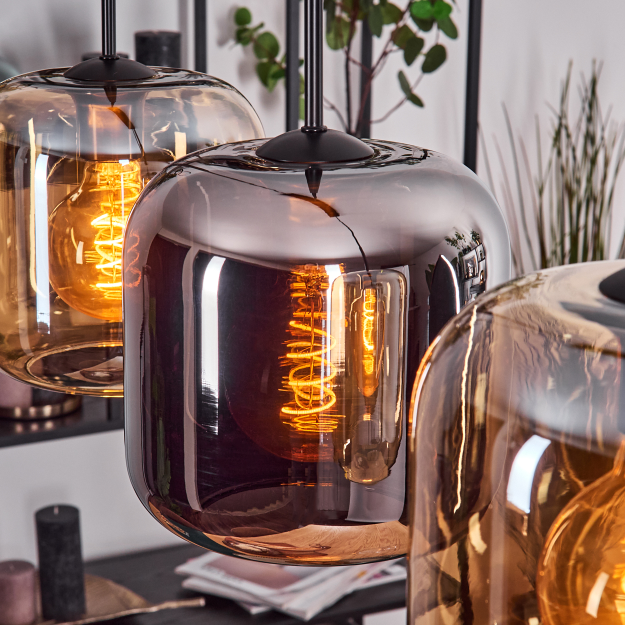 Drei Glas-Pendelleuchten mit Edison-Glühbirnen. Jeder Schirm hat eine reflektierende, rauchige Oberfläche. Hintergrund mit Dekor und Pflanzen.
