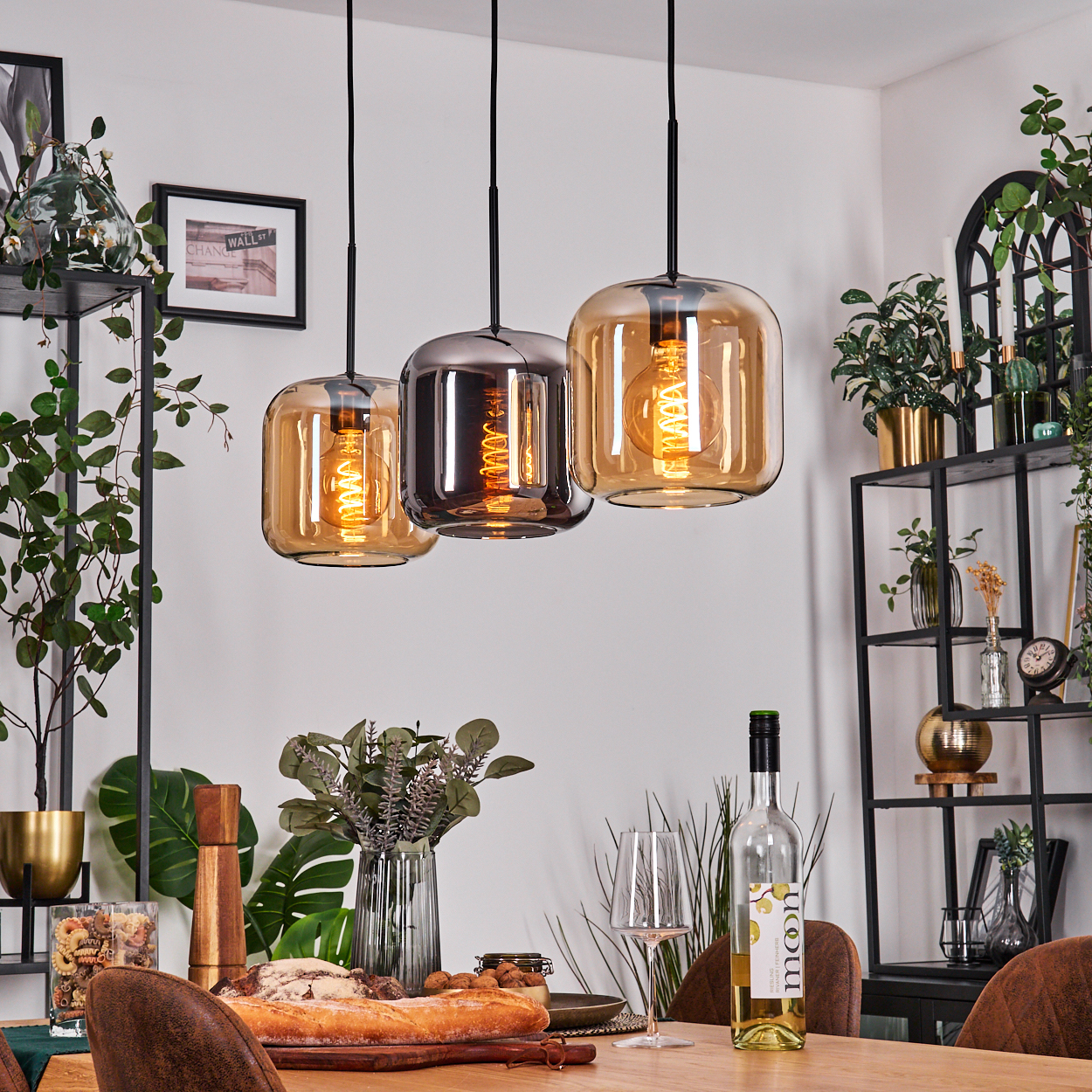 Drei Pendelleuchten über einem Tisch mit Brot, Wein und Gläsern. Regale mit Pflanzen im Hintergrund.